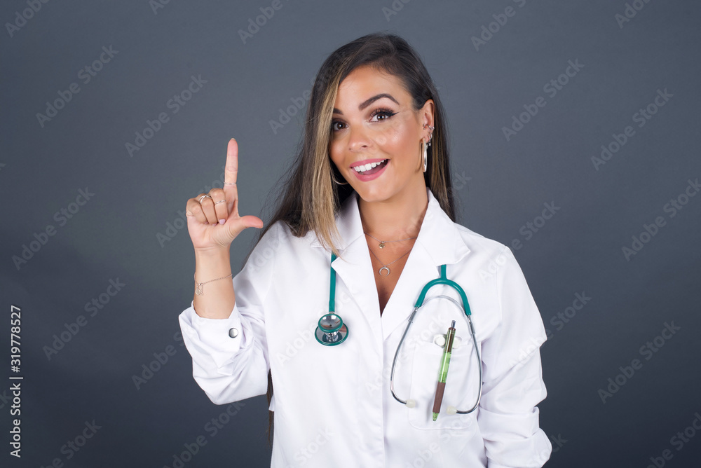 Close up portrait of pleasant looking blue eyed young doctor woman model has clever expression, raises one finger, remembers herself not to forget tell important thing. Indoors Close up portrait of pleasant looking blue eyed young doctor woman model has clever expression, raises one finger, remembers herself not to forget tell important thing. Indoors