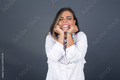 Portrait of young cute doctor woman being overwhelmed with emotions, expressing excitement and happiness with closed eyes and hands near face while smiling broadly over gray background.