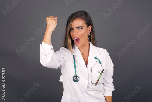 Fierce confident European dark-haired doctor woman holding fist in front of her as if is ready for fight or challenge  screaming and having aggressive expression on face.
