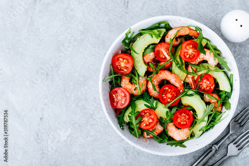 Healthy arugula salad bowl with shrimp, avocado, tomato, and sesame seeds on gray stone background.