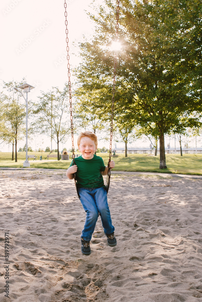 Happy smiling little preschool boy swinging on swingsat playground outside on summer day. Happy childhood lifestyle concept. Seasonal outdoor activity for kids.