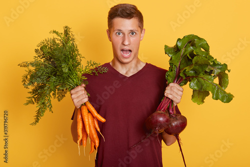 Horizontal shot of handsome man holding fresh vegetables in hands, young raw foodist, astonished posing isolated over yellow studo background with widely opened mouth. Healthy eating concept. photo