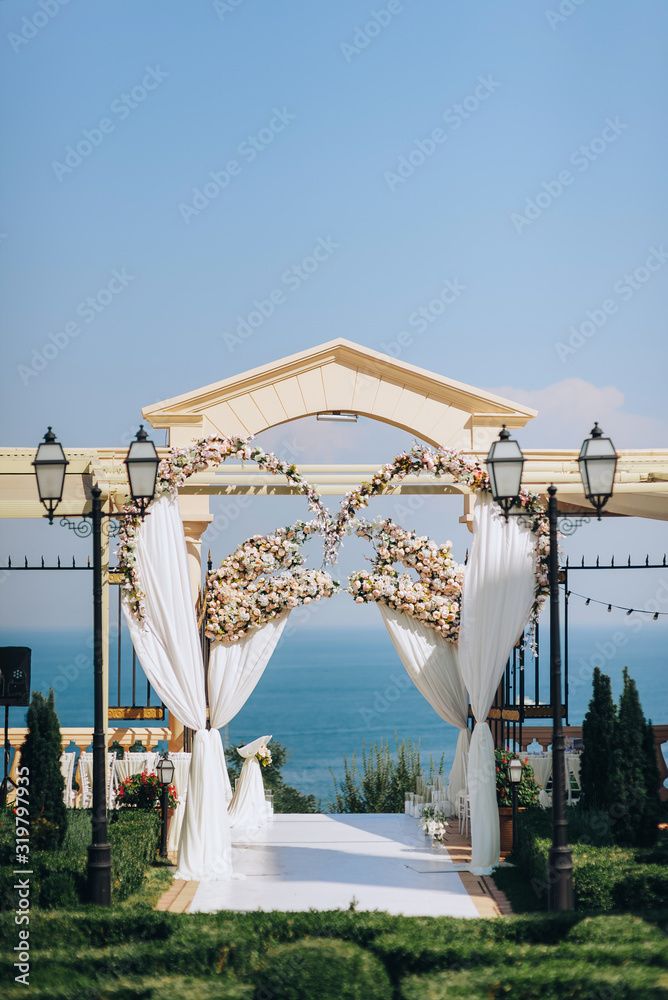 Luxurious, beautiful wedding arch with many flowers of roses near the classic columns on a background of blue sea or ocean. Summer wedding ceremony.