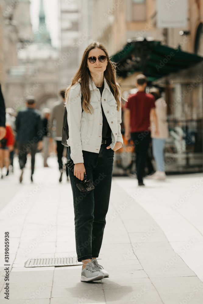 An attractive young female blogger in a white jeans jacket, black jeans, grey sneakers, and sunglasses holds the tripod with smartphone poses in the center of ancient European street full of people