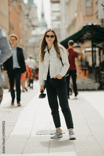 An attractive young female tourist in a white jeans jacket, black jeans, grey sneakers, and sunglasses holds the tripod with smartphone poses in the center of ancient European street full of people