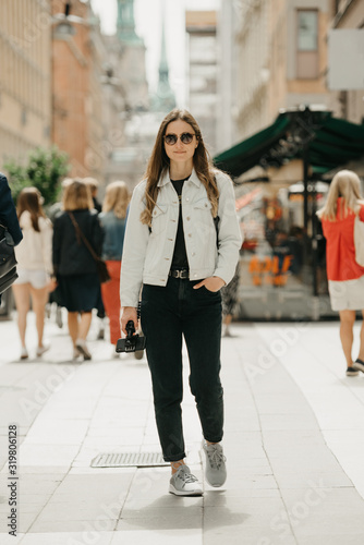 A pretty young female tourist in a white jeans jacket, black jeans, grey sneakers, and sunglasses holds the tripod with smartphone poses in the center of busy ancient European street