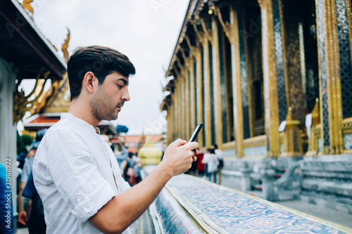 Pensive male blogger using mobile phone for making photo of asia temple visiting notable places in city, young spanish tourist enjoying exploring notable culture places taking picture on smartphone