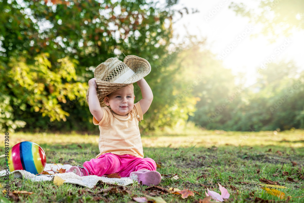 little child dresses a huge straw hat on a green lawn.