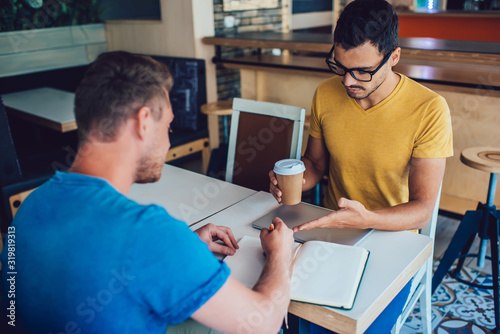 Serious male leader in casual wear and eyewear explaining strategy to employee on coffee break, skilled hipsters guys employees creating plan for cooperation on meeting writing 
