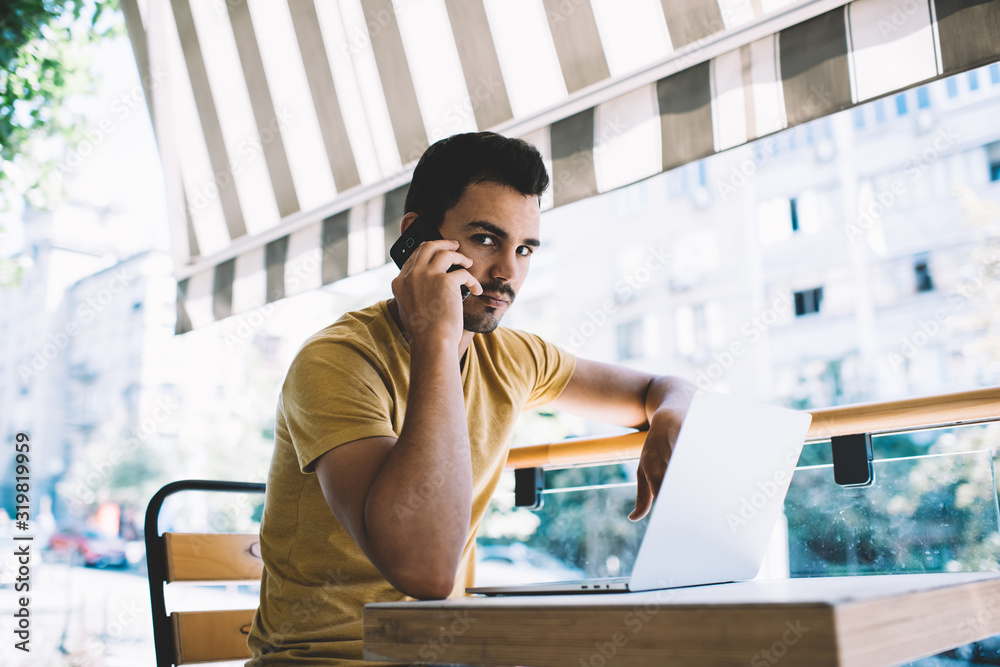 Fototapeta premium Serious caucasian male freelancer making smartphone call while working on cafe terrace remotely using laptop computer, confident man millennial concentrated on mobile phone conversation with operator.