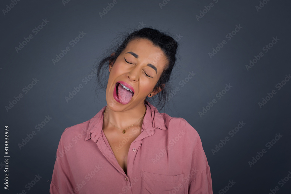 Obraz premium Beautiful caucasian woman with happy and funny face smiling and showing tongue. Wearing casual clothes and standing against gray studio background.