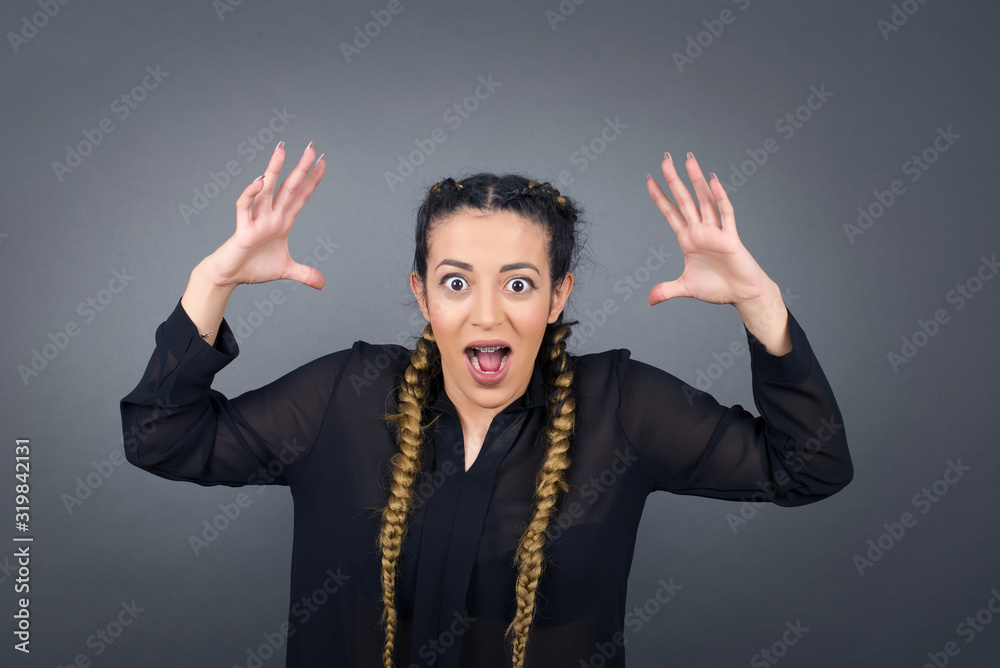 Portrait of beautiful brunette female looks with excitement at camera, keeps hands raised over head, notices something unexpected, isolated over gray wall. Lovely woman reacts on sudden news.