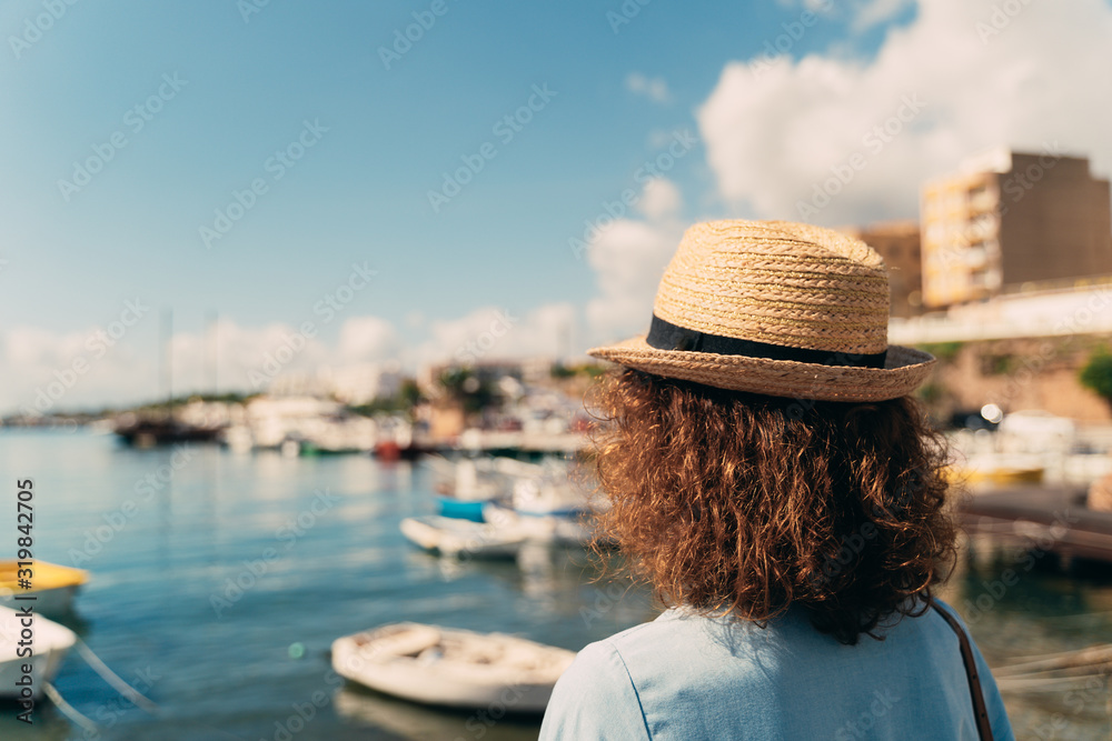 Travel tourist in blue dress and sun hat on Europe holiday looking towards a typical fishing village with white houses in the Mediterranean sea. Curly brunette woman on her 40s.