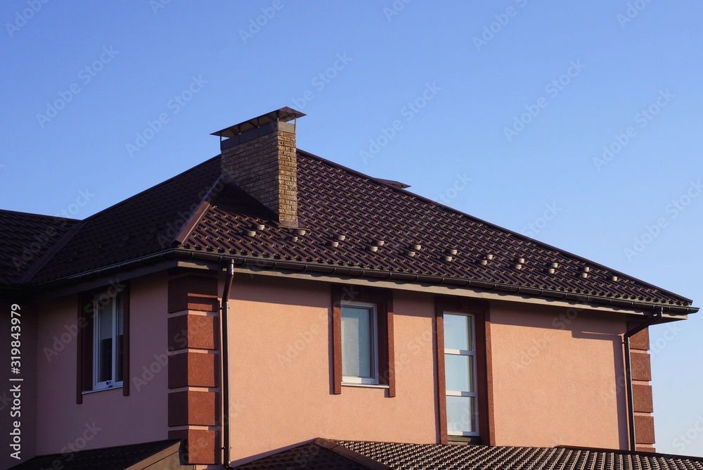 part of a brown house with windows on the wall under a tiled roof with a brick chimney against a blue sky