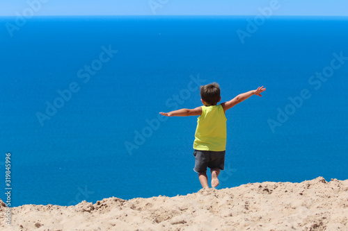 Young male caucasian preschooler jumping from a sandhill into a perfect blue sea.