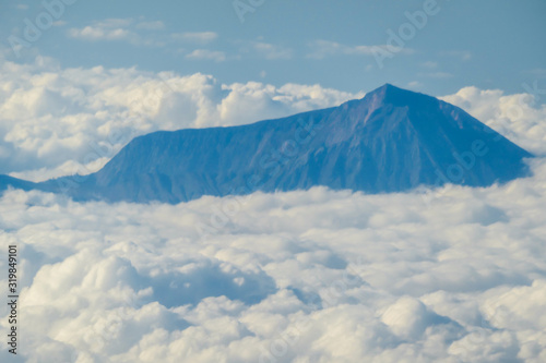 An island sneak peaking through the clouds seen from a plane s window  spotted while flying over Indonesia. Everything is covered with clouds except the mountain peak. Top down perspective.
