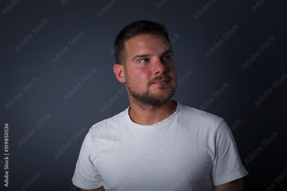 Dreamy young male with thoughtful expression, looks away keeps hands down bitting his lip thinks about something pleasant poses against gray wall.