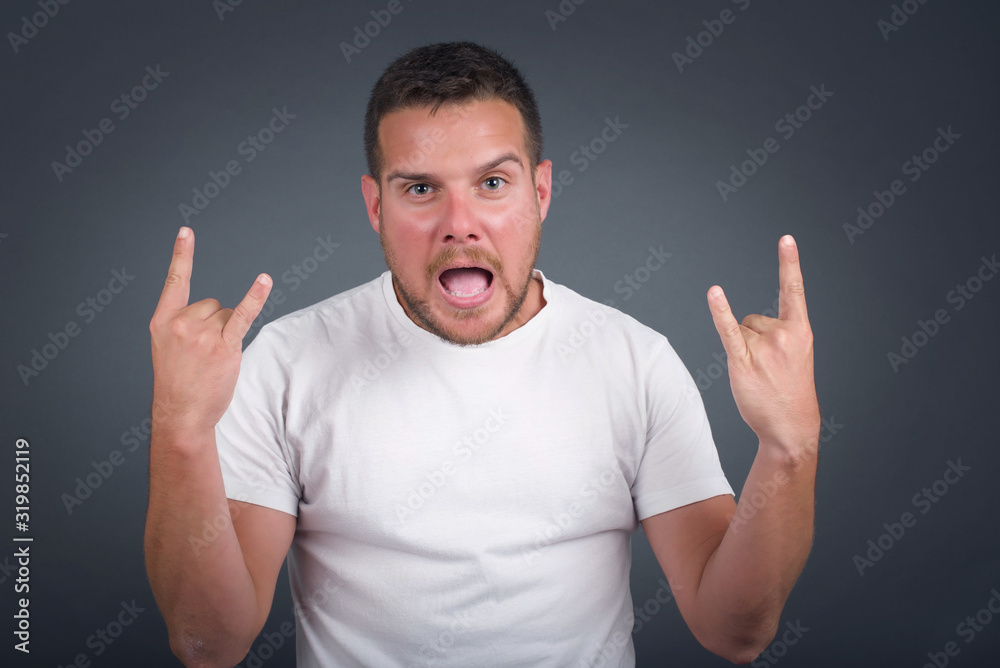 Naklejka premium Born to rock this world. Joyful man in fashionable clothes screaming out loud and showing with raised arms horns or rock gesture, expressing excitement of being on concert of band.