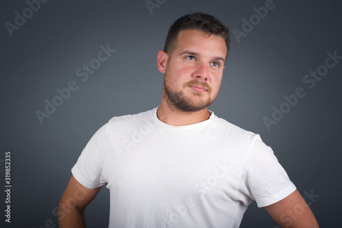 Waist up shot of joyful blue eyed boy looking up, thinking about something. Hands on hips, doubt facial expression. Standing against gray wall wondering about something happening.