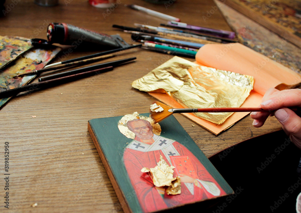 Hand with brush of gold leaf. Artist applies gold leaf to the icon. Reproduction of the icon of St. Nicholas. The process of the artist's work. Brushes and paints on the background.