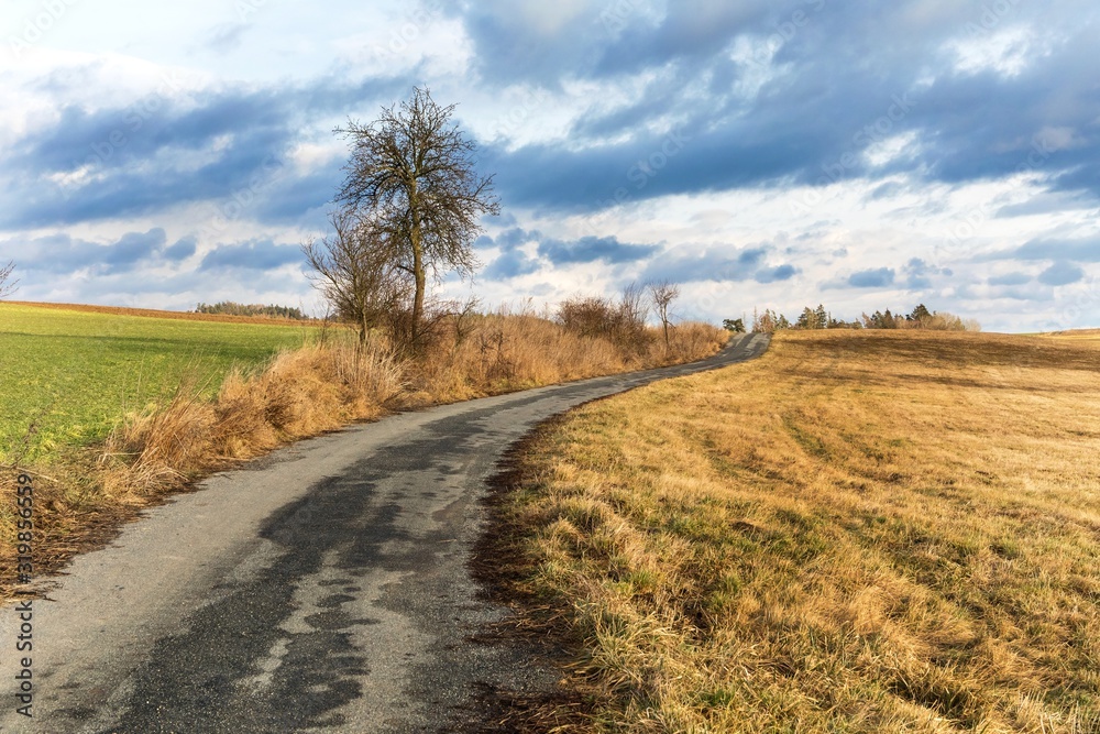 Old asphalt country road between fields. Winter time without snow. Agricultural landscape in the Czech Republic. Dramatic sky. Old asphalt country road between fields. Winter time without snow. Agricultural landscape in the Czech Republic. Dramatic sky.