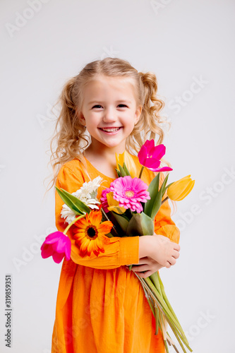 little blonde girl in an orange dress holds tulips and gerberas in her hands on a white background, a child girl smiles and holds spring flowers in her hands, space for text
