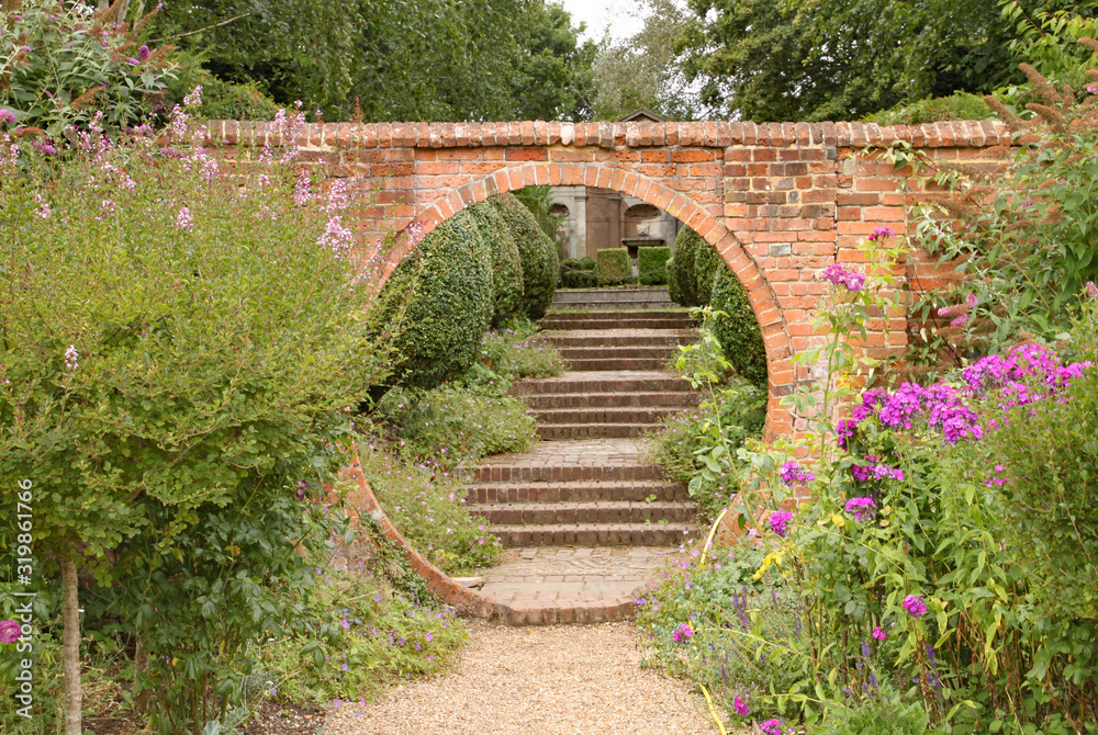 A gravel path passes through a circular archway in an old brick wall, which leads on to some old brick steps in an English country garden A gravel path passes through a circular archway in an old brick wall, which leads on to some old brick steps in an English country garden