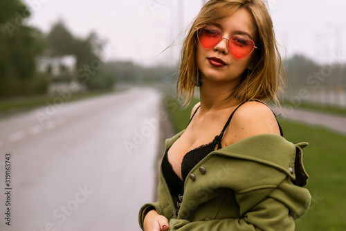 portrait of a young pretty girl in jeans and a green cloak standing during autumn little rain on the street with pink glasses  