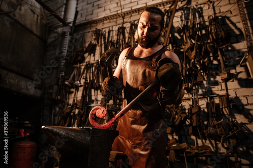 Brutal muscular artisan blacksmith standing in the workshop with a hammer, banging on a hot rod of iron on an anvil