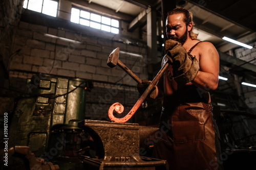 Brutal bearded man in an apron, knocking on hot metal with a hammer