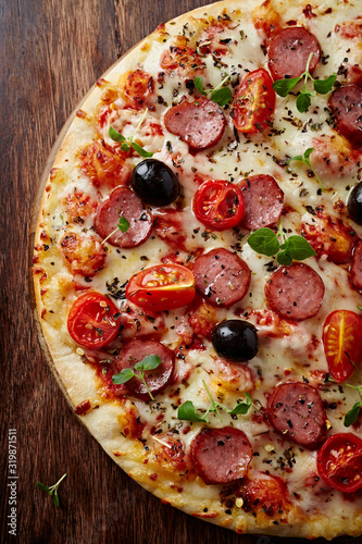 Pizza with sausage, cherry tomatoes, black olives and herbs. Home made food. Concept for a tasty and hearty meal. Brown wooden background. Top view.