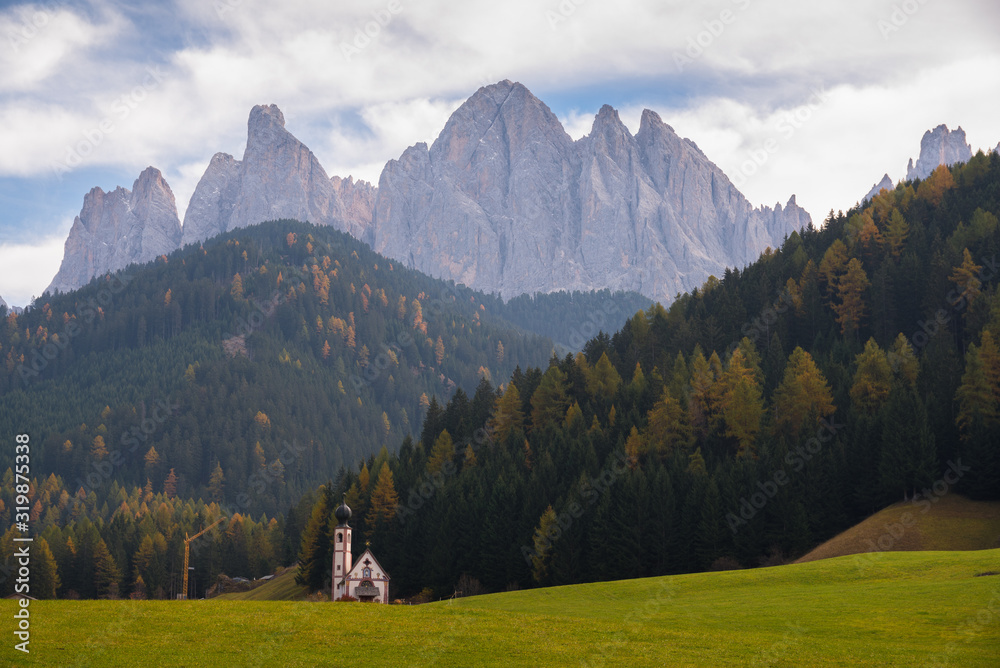 Typical scenery of small Church of San Giovanni in Ranui with dolomites Odle in background and clouds and foliage