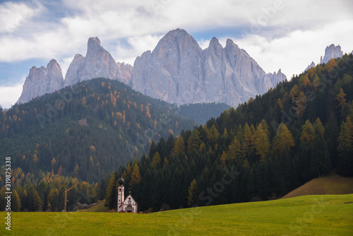 Typical scenery of small Church of San Giovanni in Ranui with dolomites Odle in background and clouds and foliage