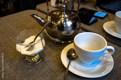 A metal teapot for tea brewing  a white ceramic cup  a glass vase for sugar on a table in a cafe.