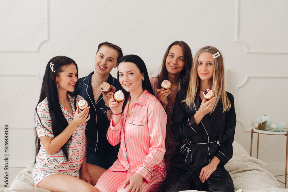 Happy pretty women in sleepwear sitting on the edge of the bed and holding cupcakes. Friendship and happiness concept Happy pretty women in sleepwear sitting on the edge of the bed and holding cupcakes. Friendship and happiness concept