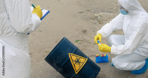 Two scientists in protective suits work near the black barrel with the sign of biological danger, one takes samples from the surface of the barrel, the other shoots the procedure on the camera.