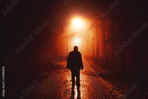 Sad man alone walking along the alley in night foggy park. Back view