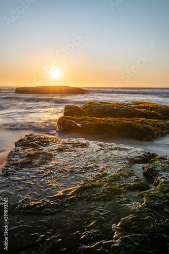 An amazing view of the sunset over the water in the Chilean coast. An idyllic beach scenery with the sunlight illuminating the green algae and rocks with orange tones and the sea in the background 