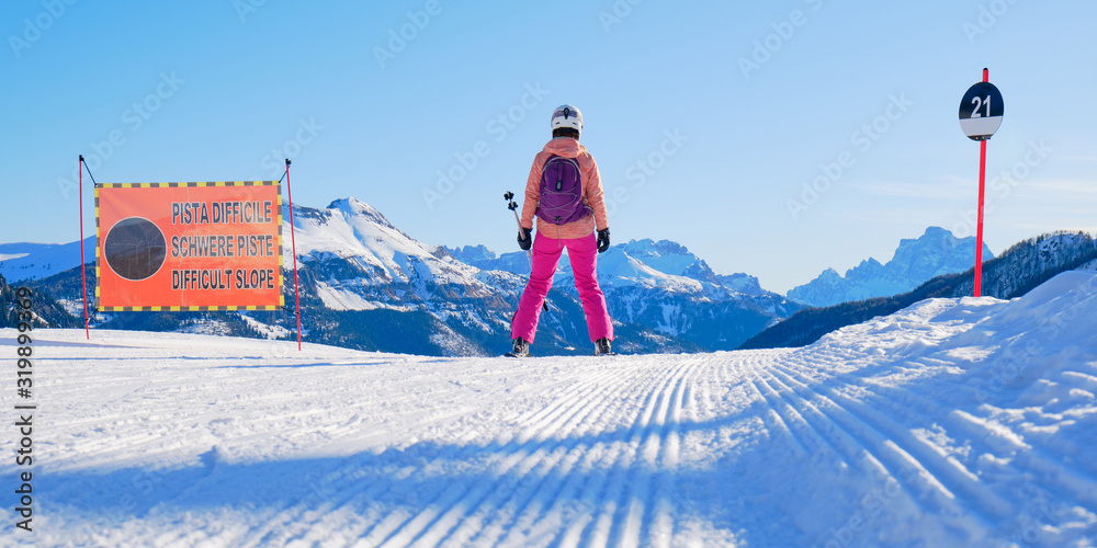 Female skier on the edge of a black ski run, looking into the distance, next to a slope sign saying Difficult Slope in Italian, German, and English, in Dolomiti Superski domain, Italy.