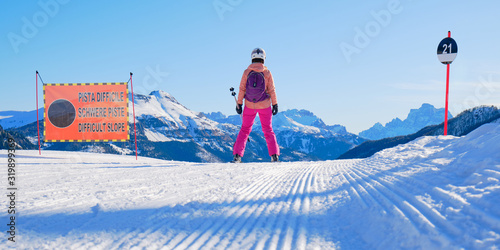 Female skier on the edge of a black ski run, looking into the distance, next to a slope sign saying Difficult Slope in Italian, German, and English, in Dolomiti Superski domain, Italy. photo