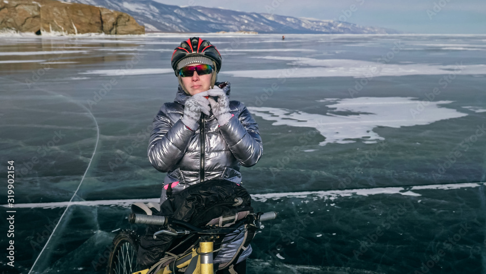 Fototapeta premium Woman stands near her bike on ice. The girl cyclist stopped to rest. The beautiful view of the sunset. The traveler is ride a cycle. Female is riding bike on the ice. Athlete puts on gear.