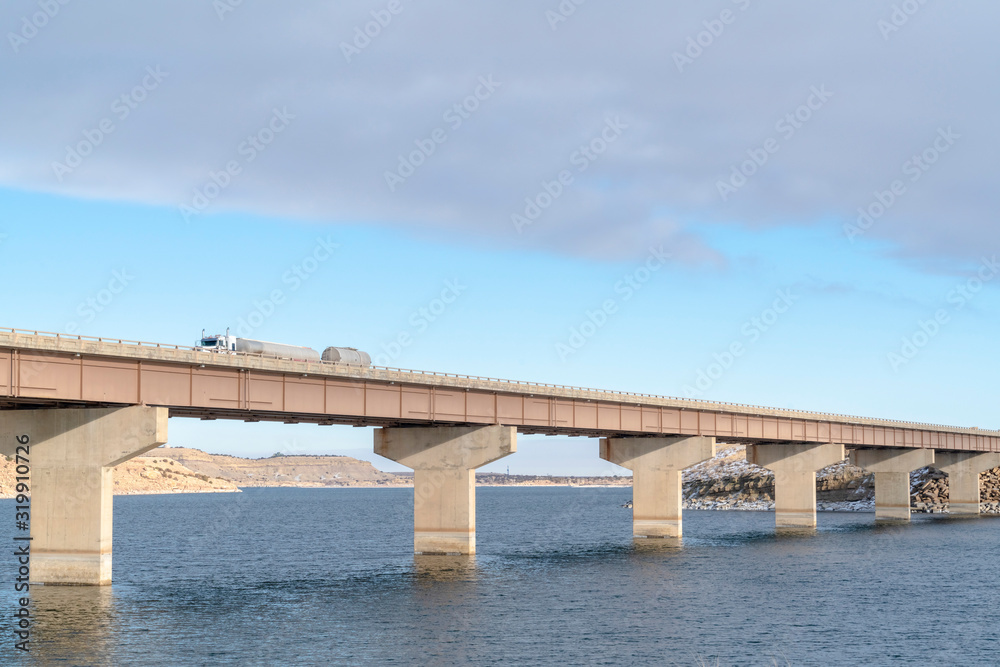 Huge truck travelling on a bridge that crosses a blue lake under cloudy sky