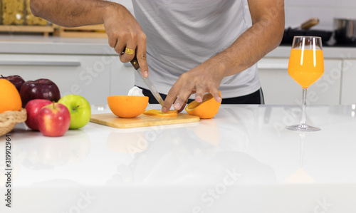 Young man sliced oranges on the wood cutting board in the kitchen, with orange juice in a beautiful glass, placed on the table as well.