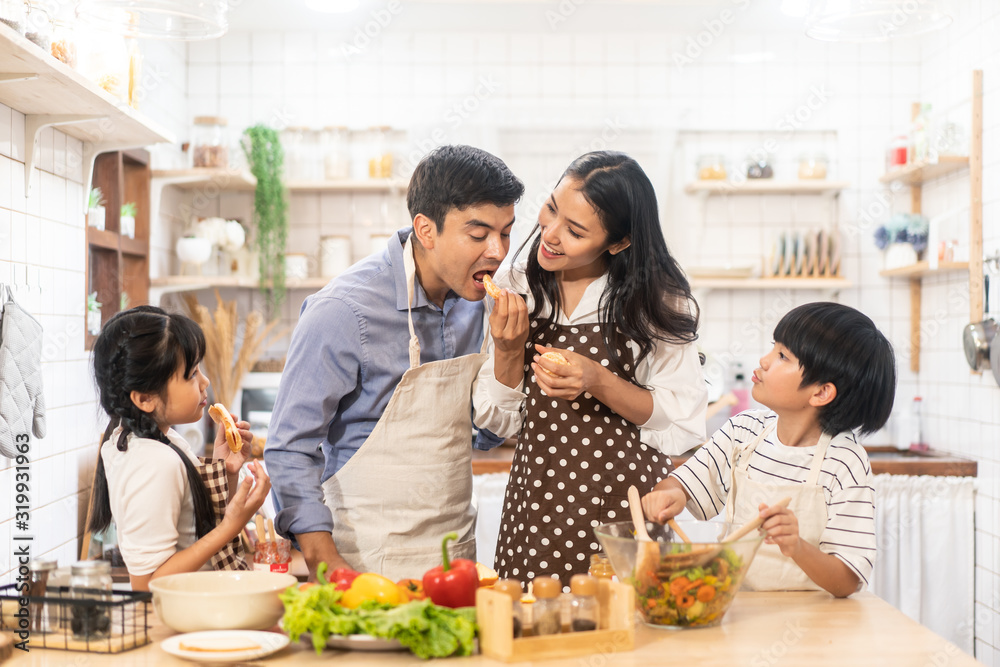 Lovely cute Asian family making food in kitchen at home. Portrait of smiling mother, dad and children standing at cooking counter. Mom feeding dad some fruit with smile. Happy family activity together