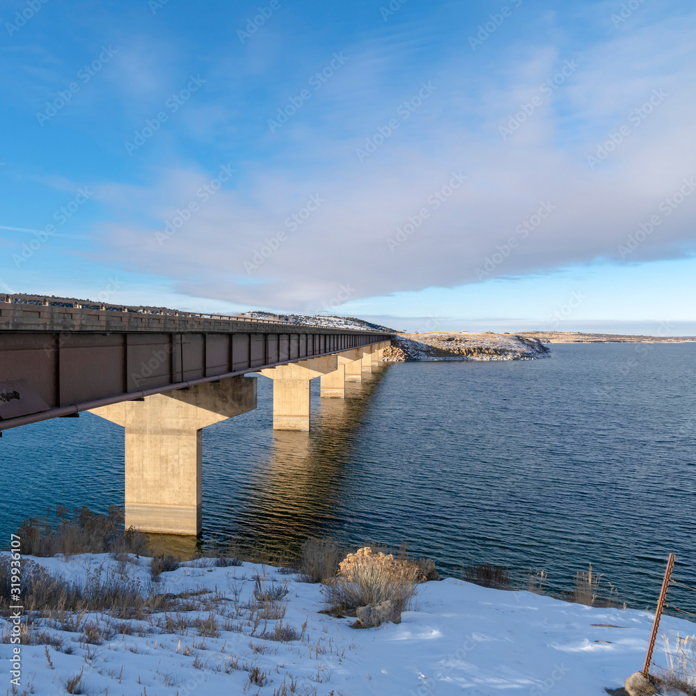 Obraz premium Square frame Bridge over a vast calm lake with snow covered shore during winter season