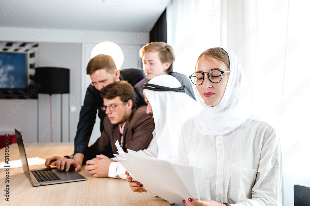 Obraz premium young girl in a national headscarf holds a computer tablet at a meeting, in the background a meeting with an Arab representative.