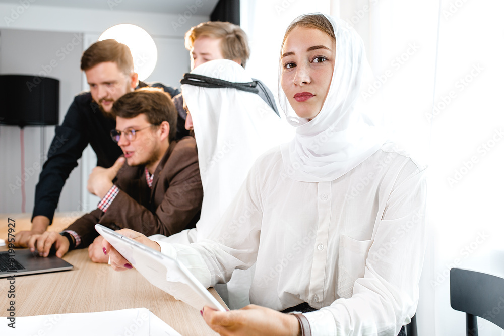 Obraz premium a young girl in a national headscarf holds a computer tablet at a meeting, in the background a meeting with an Arab representative.