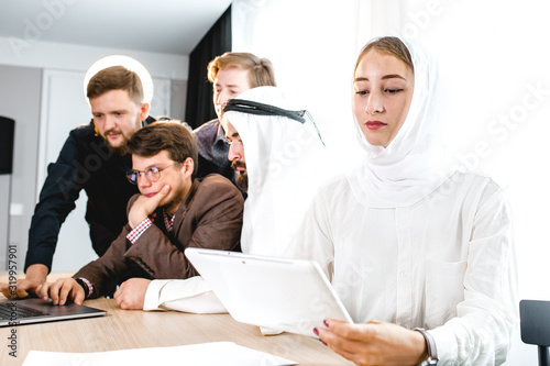 a young girl in a national headscarf holds a computer tablet at a meeting, in the background a meeting with an Arab representative.