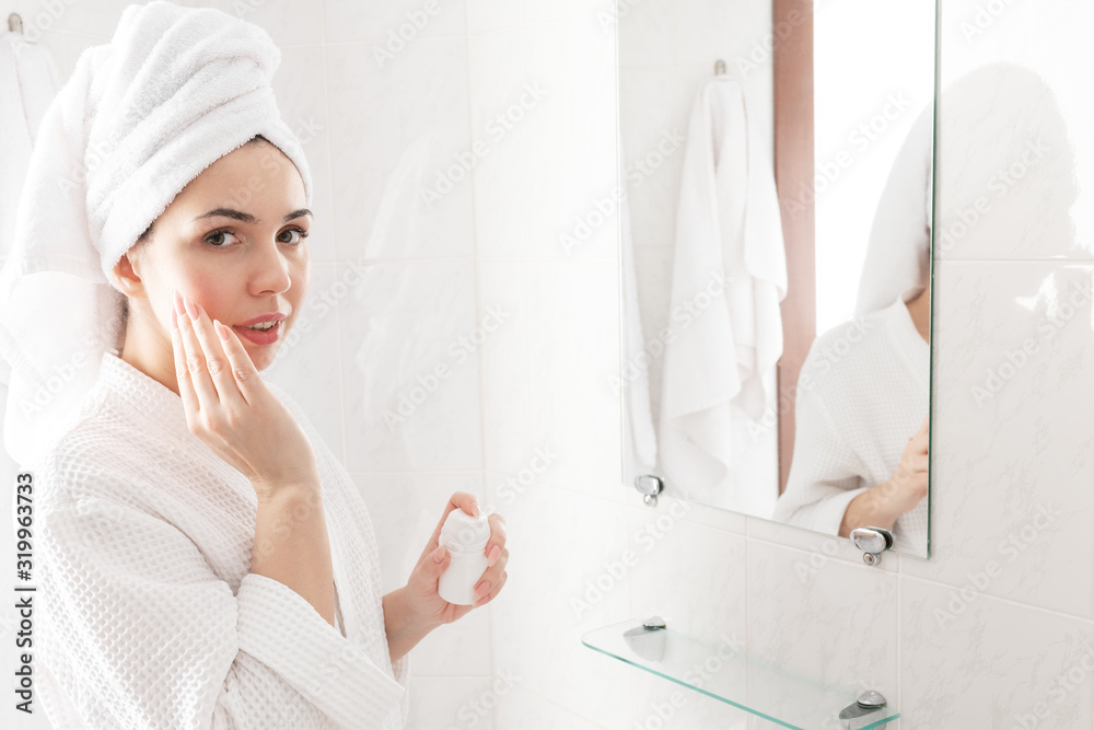 Young beautyful female with white towel on her head and in bathrobe looking at camera. Applying daily moisturizing cream. Skincare concept Young beautyful female with white towel on her head and in bathrobe looking at camera. Applying daily moisturizing cream. Skincare concept