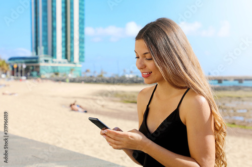 Smiling young woman in tank top using mobile phone and texting on seaside town in summer